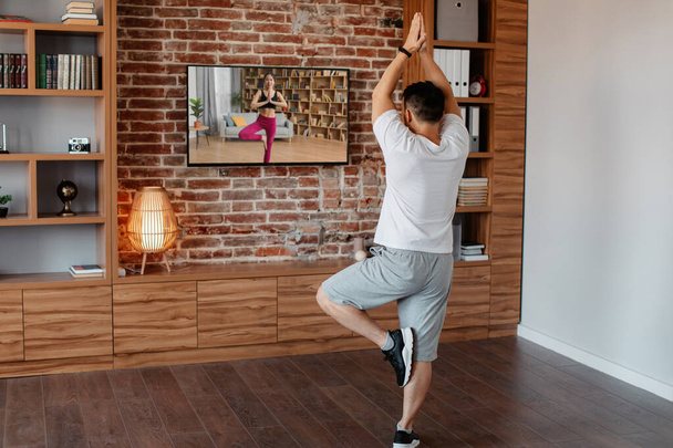 Back view of mature man doing yoga while watching video training on TV, standing in tree pose with arms raised, exercising at home. Meditation and relaxation, sporty lifestyle - Foto, Imagem