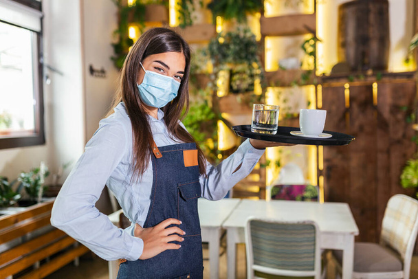 Female waitress in a medical protective mask serves the coffee in restaurant durin coronavirus pandemic representing new normal concept - Valokuva, kuva