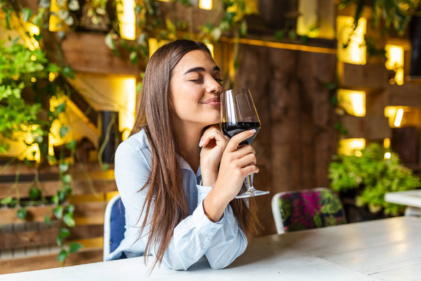Beautiful woman tasting wine while sitting in restaurant. Image of cute pretty young woman sitting in cafe holding glass and drinking wine. portrait of a beautiful wine tasting tourist woman. - 写真・画像