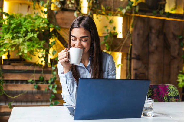 Young business woman in sitting at table in cafe and drinking coffee. On table is laptop and smartphone Freelancer working in coffee shop. Student learning online. - Fotó, kép