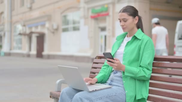 Молодая женщина с помощью смартфона и ноутбука, сидя на открытом воздухе на скамейке - Кадры, видео