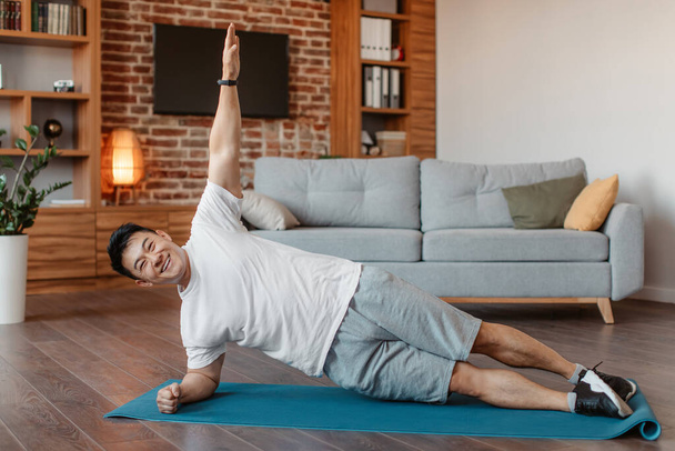 Happy asian middle aged man in sportswear doing side plank on floor mat in living room interior, copy space. Sport at home, workout, fitness and exercises, active lifestyle - Φωτογραφία, εικόνα
