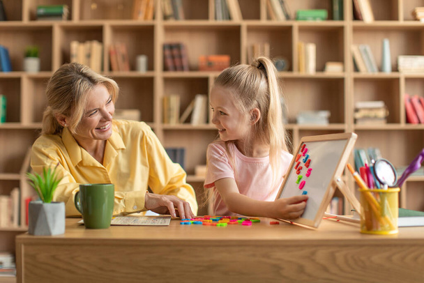 Preschool education. Happy professional female teacher exercising with pretty girl, cute child reading English letters and making words on whiteboard - Foto, Imagem