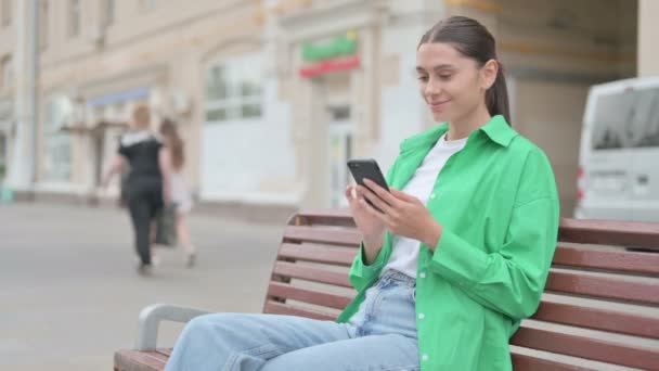 Γυναίκα Γιορτάζοντας σε απευθείας σύνδεση επιτυχία στο Smartphone, ενώ κάθεται σε εξωτερικούς χώρους για Bench - Πλάνα, βίντεο