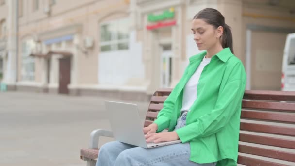 Απασχολημένη νεαρή γυναίκα που χρησιμοποιεί φορητό υπολογιστή κάθεται σε εξωτερικό χώρο στο Bench - Πλάνα, βίντεο