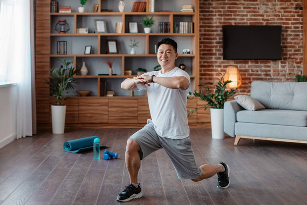 Athletic asian mature man making lunge, doing exercises, working out legs muscles at home and smiling at camera. Middle aged korean male practicing domestic sports - Foto, imagen