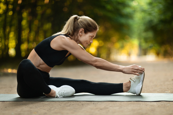 Sporty blonde woman stretching on yoga mat at public park, active middle aged lady having workout outdoors, enjoying morning activity, side view, copy space. Open air fitness concept - Foto, imagen