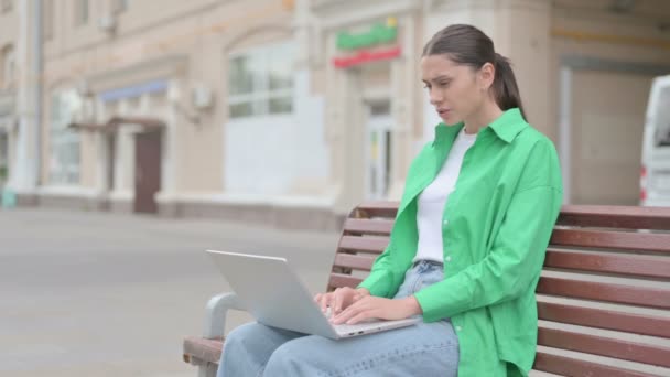 Молодая женщина реагирует на потери на ноутбуке, сидя на открытом воздухе на скамейке - Кадры, видео