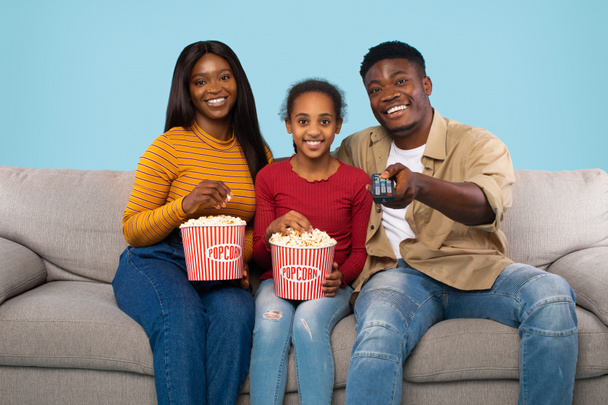 Weekend leisure. Black family watching TV and eating popcorn, sitting on couch over blue studio background. Man switching channels with remote control - 写真・画像
