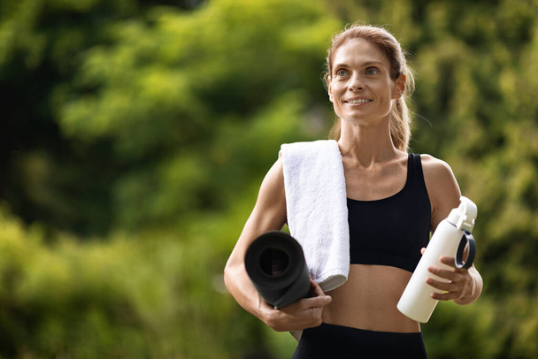 Happy slim lady in black sportswear having yoga at public park, holding towel, fitness mat and bottle of water, having break while training, panorama with copy space. Fitness outdoos concept - Фото, зображення