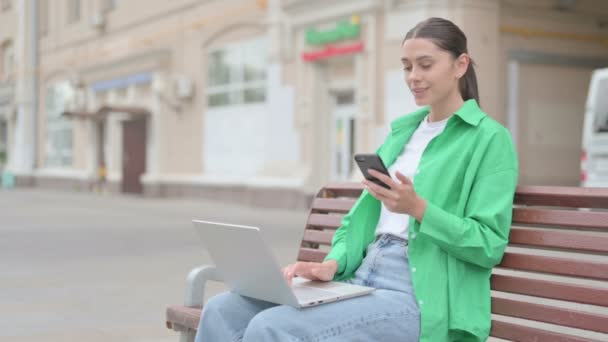 Молодая женщина разговаривает по телефону и с ноутбуком, сидя на открытом воздухе на скамейке - Кадры, видео