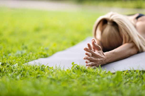 Closeup shot of unrecognizable blonde lady lying on yoga mat with hands crossed in public park, athletic woman having yoga practice outdoors in summer, panorama with copy space - Фото, изображение