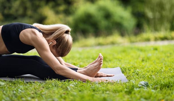 Side view of slim well-fit blonde lady sitting on fitness mat, reaching her knees with forehead, stretching her body in public park, doing yoga outdoors, panorama with copy space - Foto, Imagen