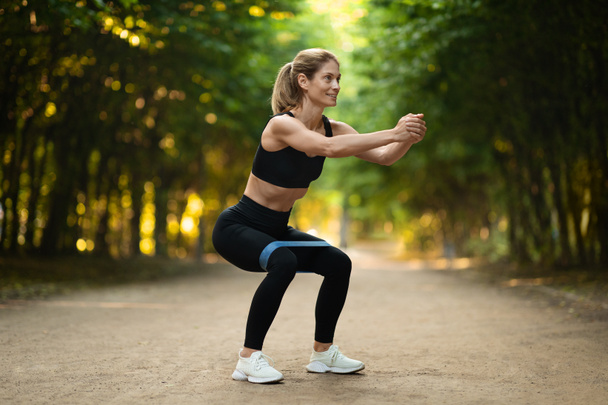Motivated blonde woman in black sportswear and white sneakers exercising with resistance band outdoors, looking at copy space and smiling, full length shot. Fitness equipment concept - Φωτογραφία, εικόνα
