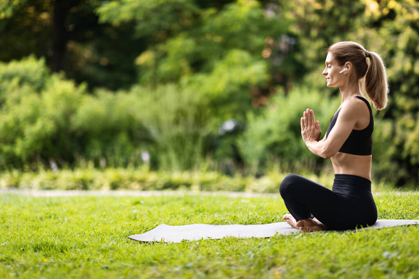 Peaceful well-fit blonde woman in black tight sportswear sitting on fitness mat in lotus position with closed eyes, using wireless earbuds, meditating alone at public park, side view, copy space - Photo, image