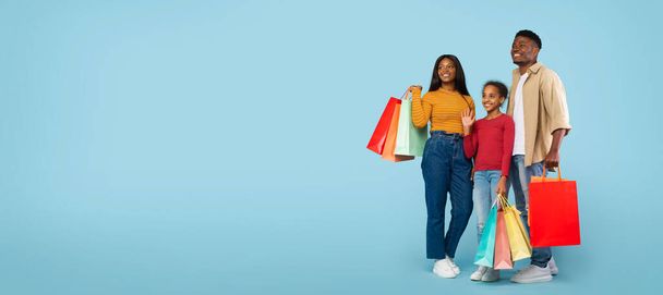 Family shopping concept. Happy african american parents and their daughter carrying colorful shopper bags over blue background, panorama with free space - Foto, imagen