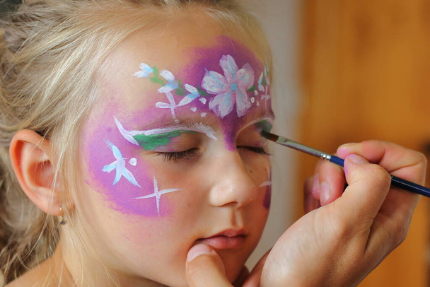 Children's face painting. The artist paints a princess crown on a small white blonde girl. - Fotó, kép