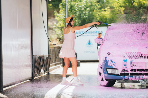 Brunette with long hair from a high-pressure hose applies a cleaner on the car at a car wash - Photo, Image