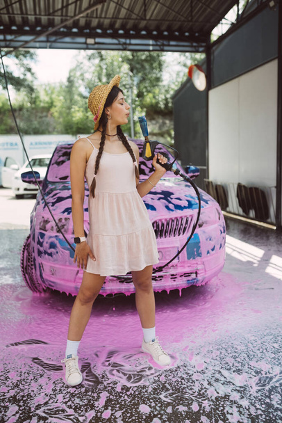 Mladá žena vysokotlaká hadice stojí před autem pokryté růžovou pěnou - Fotografie, Obrázek