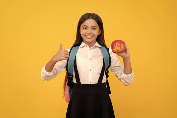 Niño feliz niña dar pulgares hacia arriba la celebración de la manzana y el bolso de la escuela fondo amarillo, la educación. - Foto, Imagen
