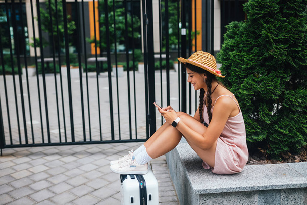 Junge schlanke Frau sitzt auf einer Bank mit Handy, grauer Koffer neben den Beinen, Seitenfoto - Foto, Bild