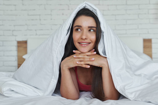 Dreamful young woman looking out of a blanket while lying in bed at home - Фото, изображение