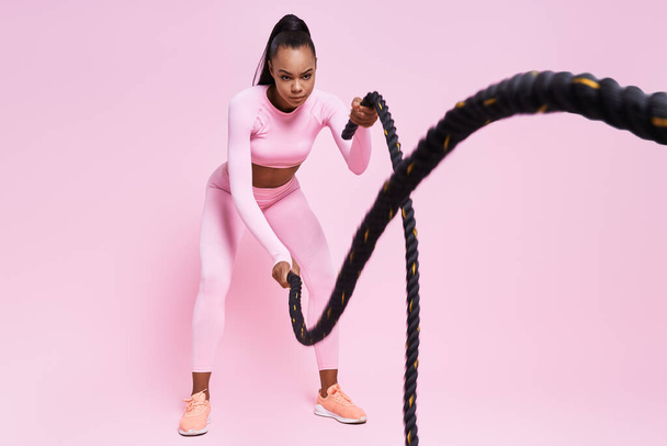 Confident young African woman exercising with battle ropes against pink background - Foto, Imagem