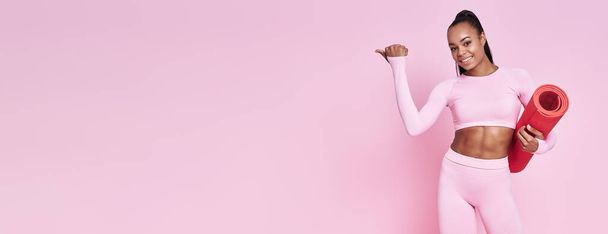 Happy woman in sports clothing carrying exercise mat and pointing away against pink background - 写真・画像