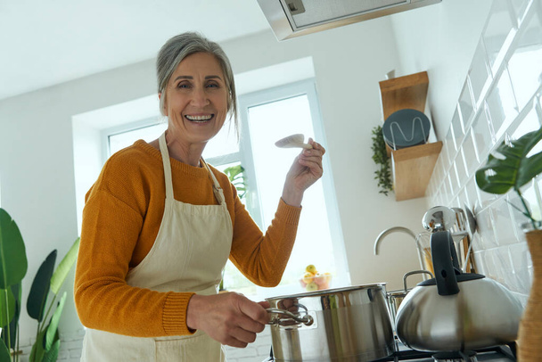 Happy senior woman looking at camera while cooking at the domestic kitchen - Photo, image