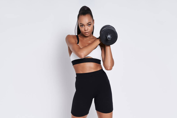 Confident young African woman holding dumbbell while standing against white background - Foto, Imagem