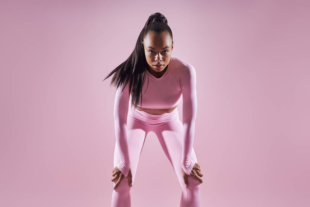 Concentrated African woman in sports clothing holding hands on knees against pink background - Foto, Imagem