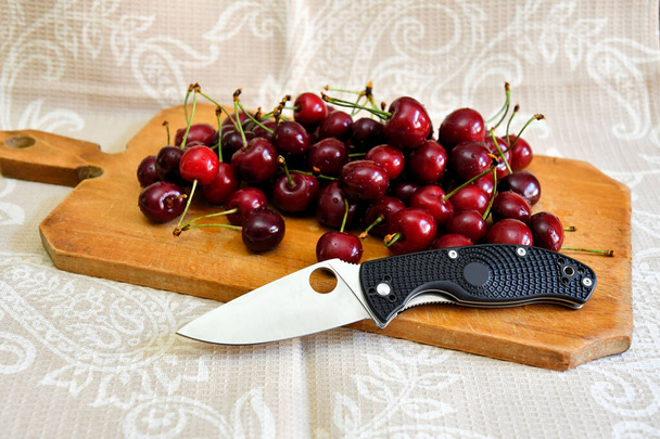 Folding knife cutting edge black handle fresh red sweet cherry vegetarian natural product brown wooden plate background - Fotoğraf, Görsel