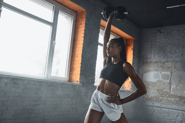 Confident young African woman using dumbbell while exercising in gym - Foto, Imagen