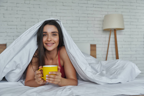 Beautiful woman looking out of a blanket and holding coffee cup while lying in bed at home - Fotografie, Obrázek