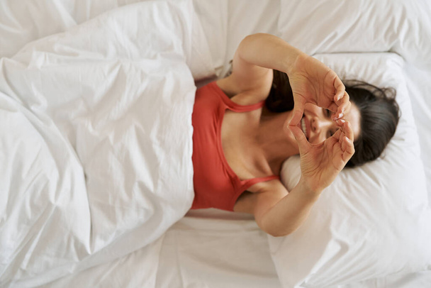 Top view of beautiful young woman gesturing heart shape and smiling while lying in bed - Fotó, kép