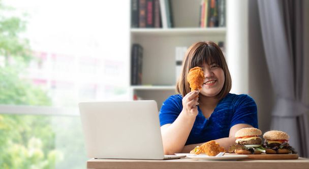 Mujer hambrienta con sobrepeso sosteniendo pollo frito, hamburguesa en un plato de madera y pizza en la mesa, durante el trabajo desde casa, subir de peso problema. Concepto de trastorno de atracones (BED)). - Foto, imagen