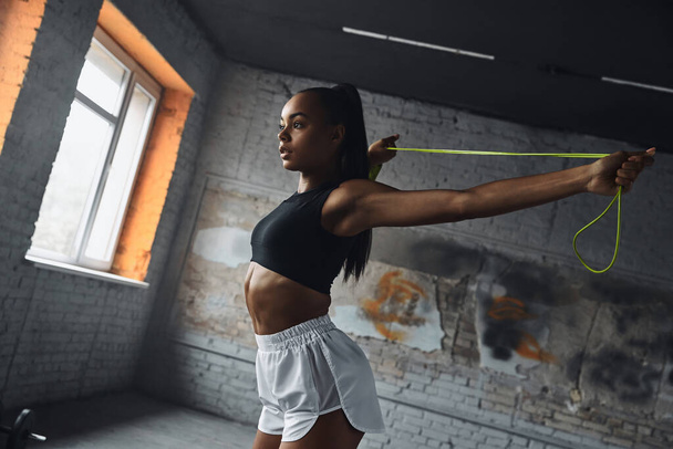 Confident young African woman using jumping rope while doing stretching exercises in gym - Фото, зображення