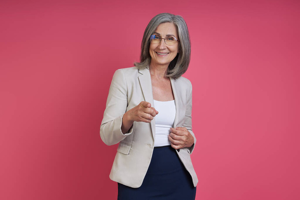 Happy senior woman in formalwear pointing camera while standing against pink background - Fotó, kép