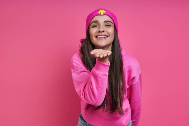 Beautiful young woman in funky hat blowing a kiss against pink background - Photo, image