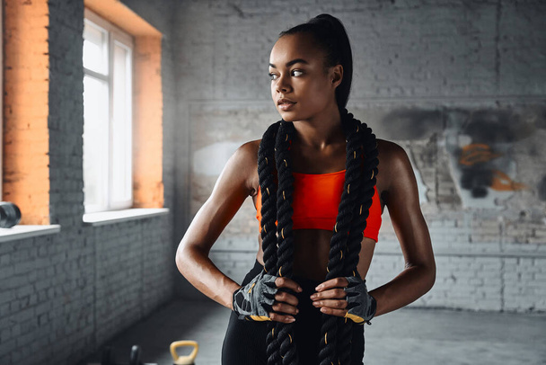 Attractive young African woman carrying battle rope on shoulders while standing in gym - Photo, Image