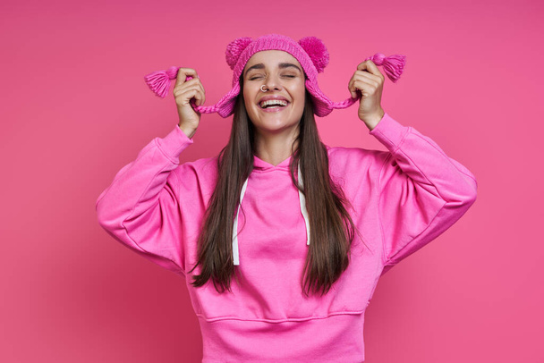 Happy young woman in hooded shirt playing with her funky hat against pink background - Foto, afbeelding