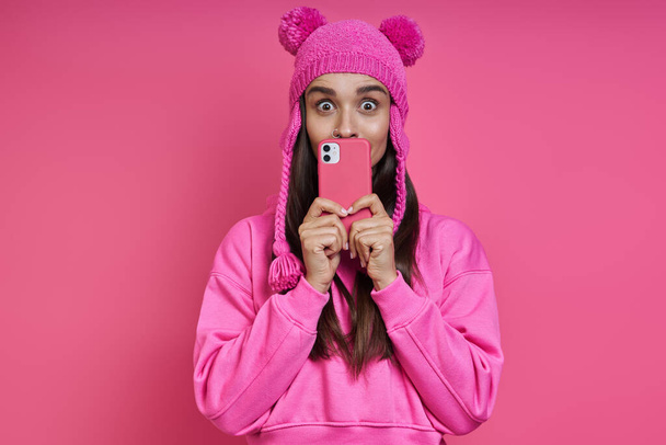 Surprised young woman in funky hat covering mouth with smart phone while standing against pink background - Valokuva, kuva