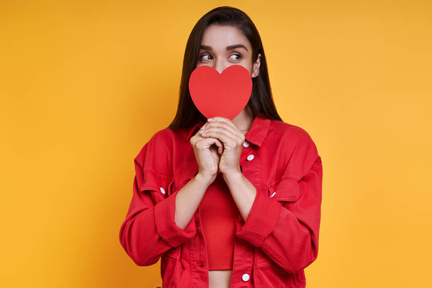 Beautiful young woman covering mouth with paper heart with hand against yellow background - Φωτογραφία, εικόνα