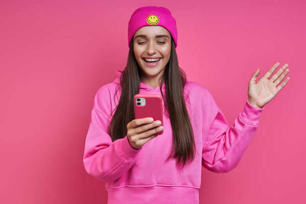 Beautiful young woman in hooded shirt and funky hat holding smart phone against pink background - Zdjęcie, obraz