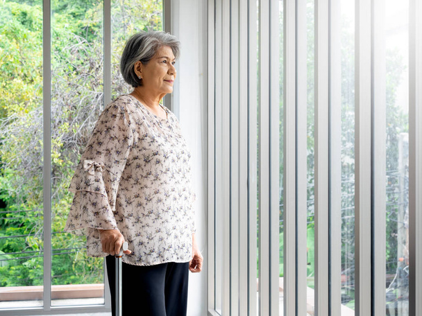 Asian senior woman, white hair standing with a cane and looking out the glass window, indoors with copy space. Elderly lady patient using walking cane. Strong health, medical care concepts. - Photo, Image