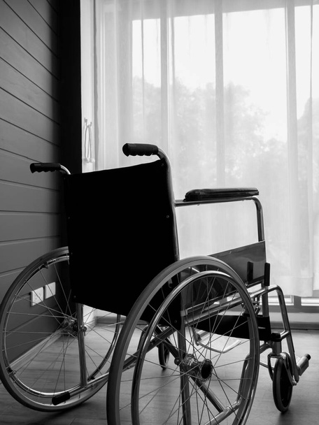 Вид ззаду на порожнє чорне інвалідне крісло біля стіни з білою завісою у скляному вікні у вертикальному стилі вітальні, що чекає на послуги пацієнта. Самотній інвалідний візок з ніким у лікарняній кімнаті
. - Фото, зображення