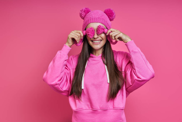 Joyful young woman in hooded shirt playing with her funky hat against pink background - Foto, Imagen