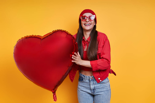 Happy young woman holding heart shape balloon against yellow background - Fotó, kép