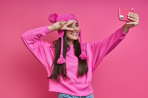 Beautiful young woman in funky hat making selfie and gesturing against pink background - Fotografie, Obrázek