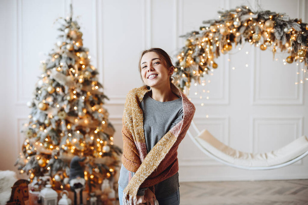 Счастливая женщина в сером свитере и шарфе стоит дома на фоне рождественской елки. Удовлетворенный декоратор на фоне рождественского интерьера. - Фото, изображение